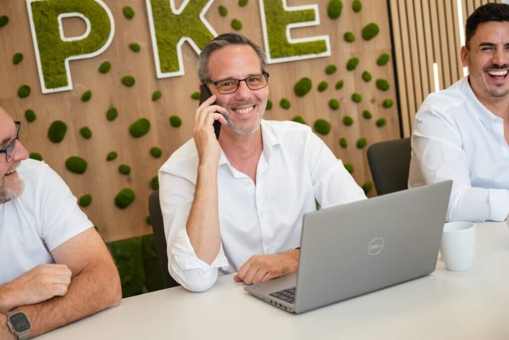 Mann am Schreibtisch telefoniert fröhlich mit Laptop, moderner Bürohintergrund mit grüner Wanddekoration.